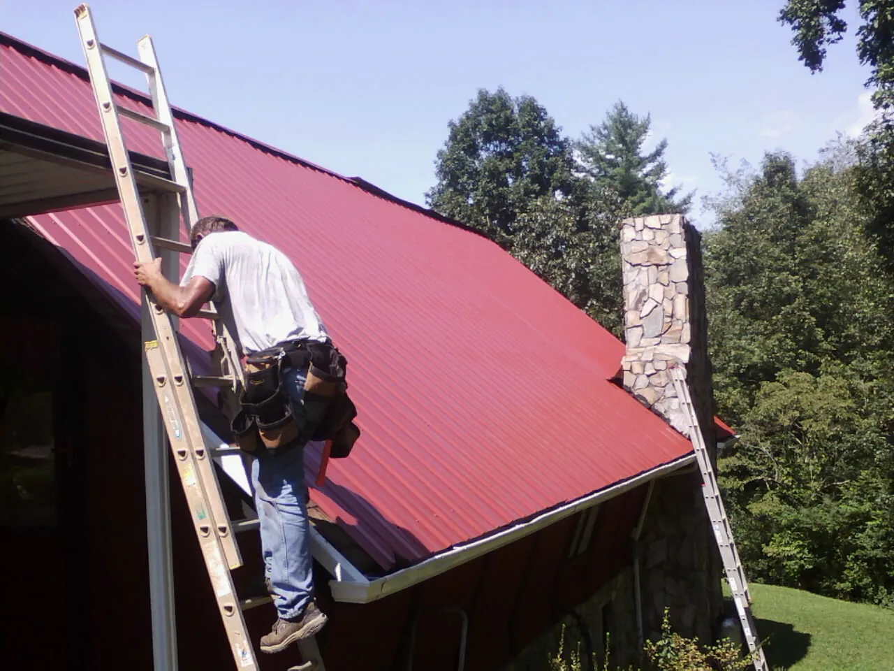 Roofer on a ladder beside a red metal roof and stone chimney — Soffit & Fascia Repair by Roofing Brothers in Elmore